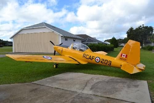 A yellow aircraft with RCAF markings is parked on a concrete pad in front of a house.
