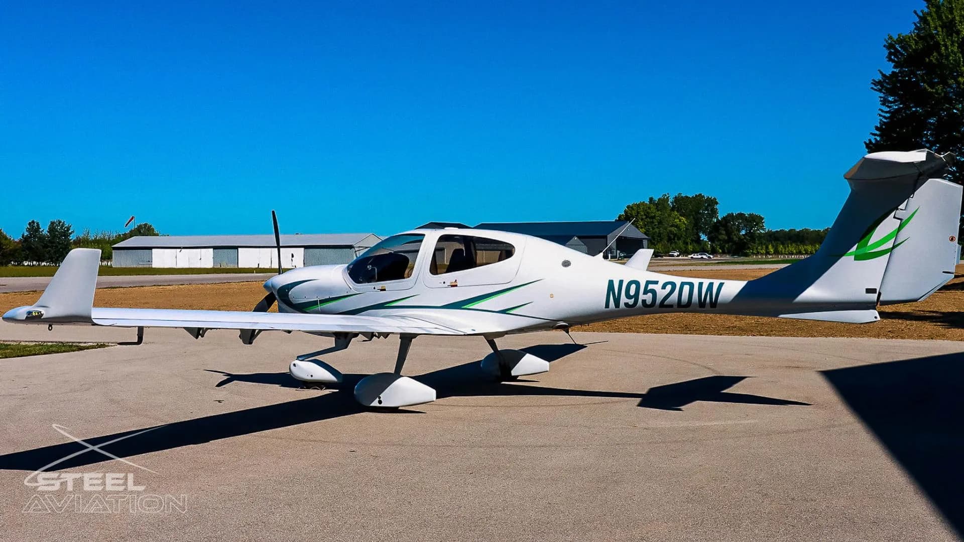 White Diamond DA40 aircraft with green accents and registration N952DW parked on an airfield with hangars in the background.