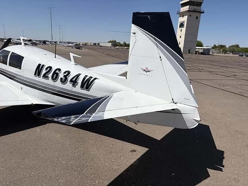 The tail section of the white Mooney M20 aircraft N2634W is visible on an airfield, with an air traffic control tower in the background.