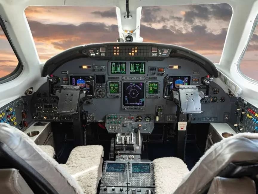 The cockpit of a business jet, showing a complex array of digital displays, instruments, and controls, with a sunset sky visible through the windshield.
