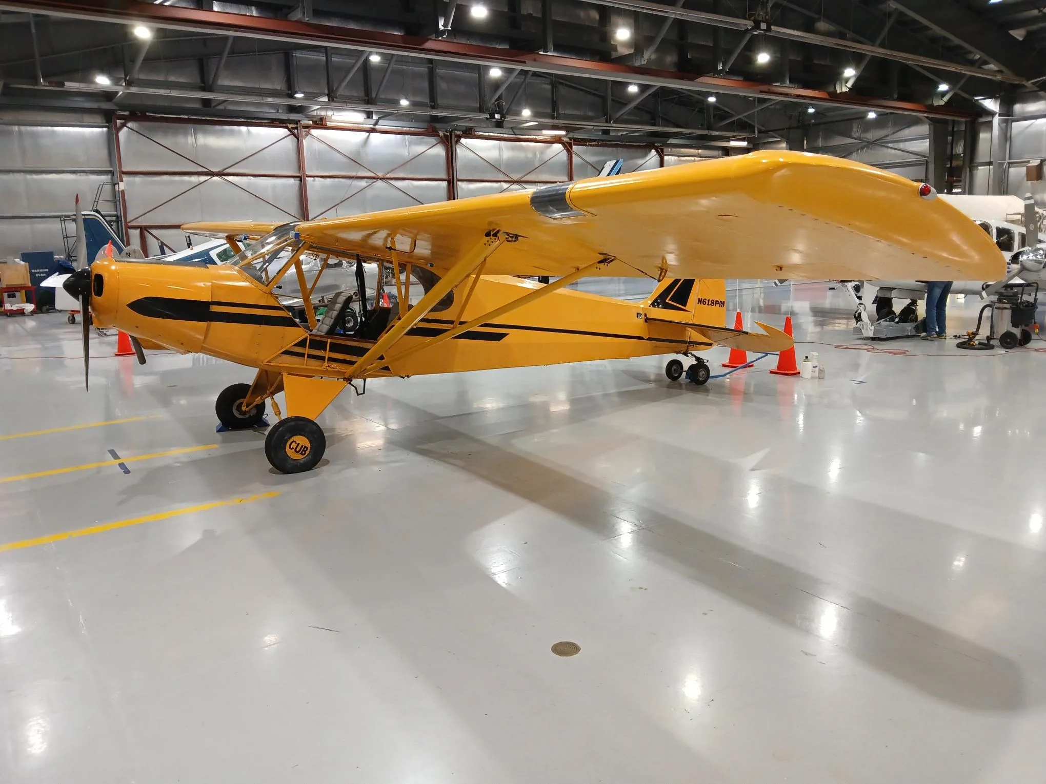 A yellow Piper Cub-style aircraft, N618PM, is seen from a different angle inside a hangar.