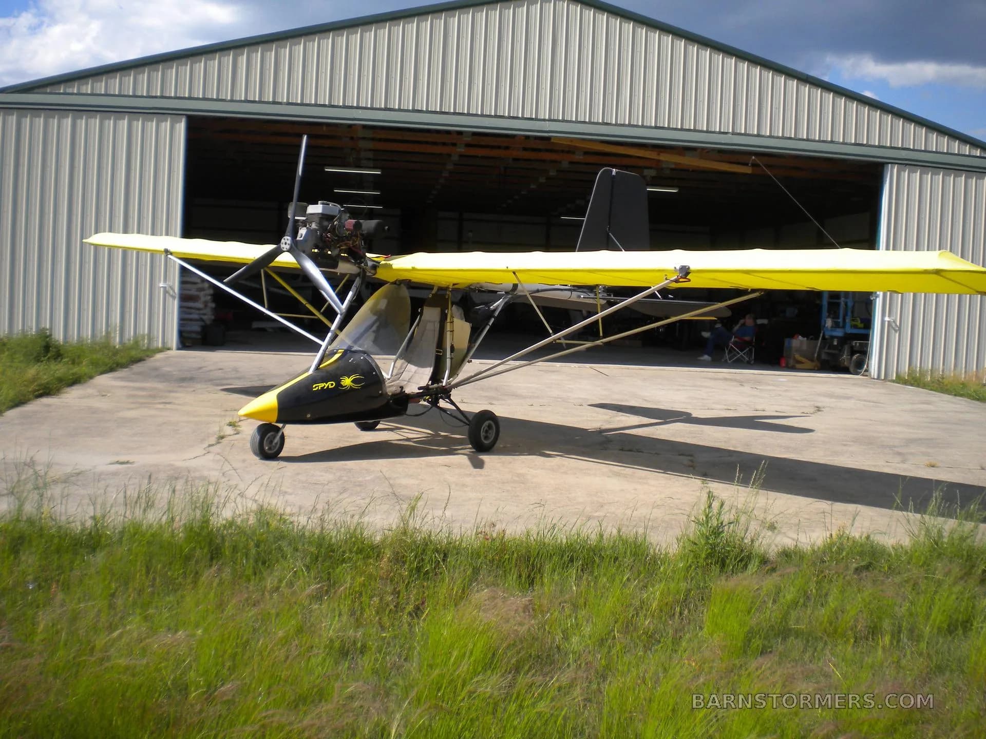 A yellow-winged ultralight aircraft with a black nose cone, parked on a concrete pad in front of an open hangar.