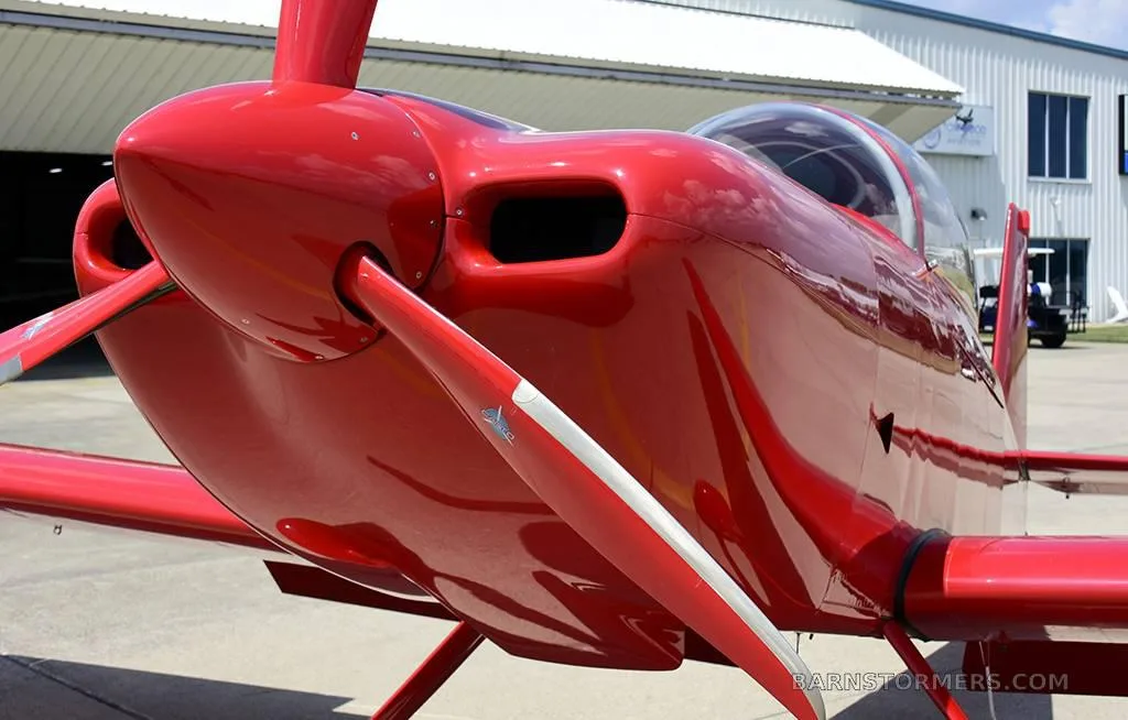 A close-up front view of a red single-engine aircraft, highlighting its propeller, engine cowling, and canopy.