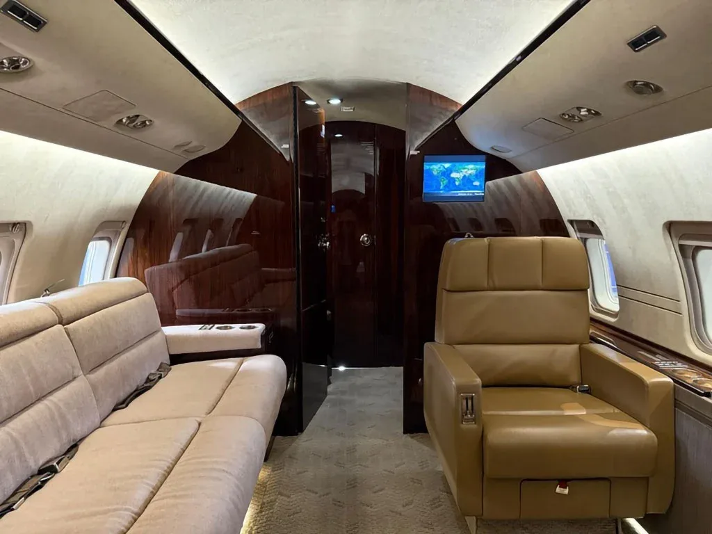 The private jet cabin features a beige sofa and a matching single seat, with dark wood paneling and a screen displaying a map.