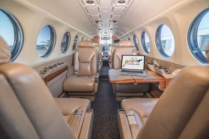 The aircraft cabin features tan leather seats, a small table with a laptop, and round windows.