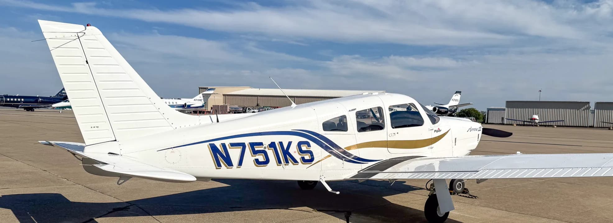 A white Piper Arrow III aircraft with blue and gold stripes, tail number N751KS, parked on an airport tarmac with other aircraft in the distance.