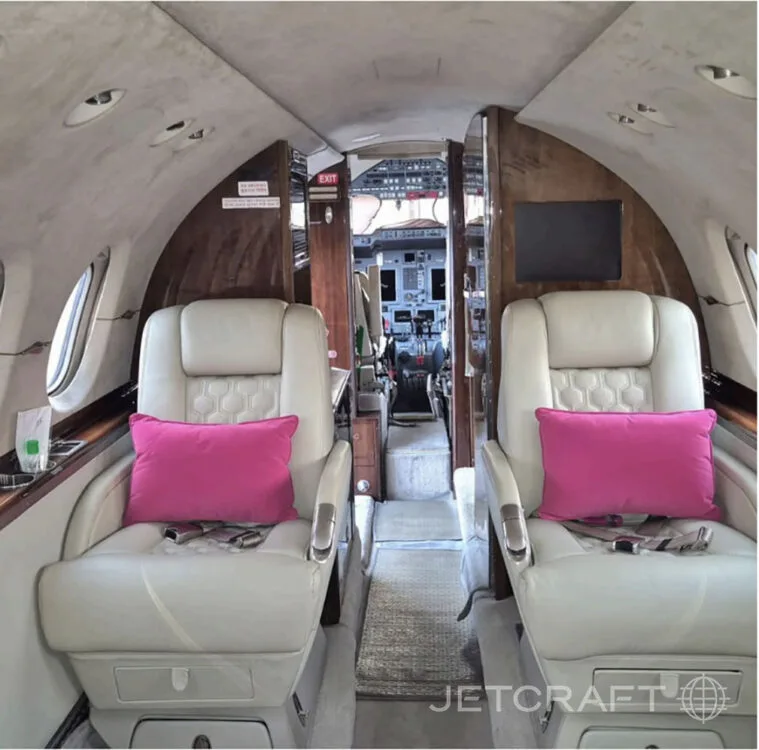 The interior cabin of a business jet with two cream-colored seats and pink pillows, looking towards the cockpit.