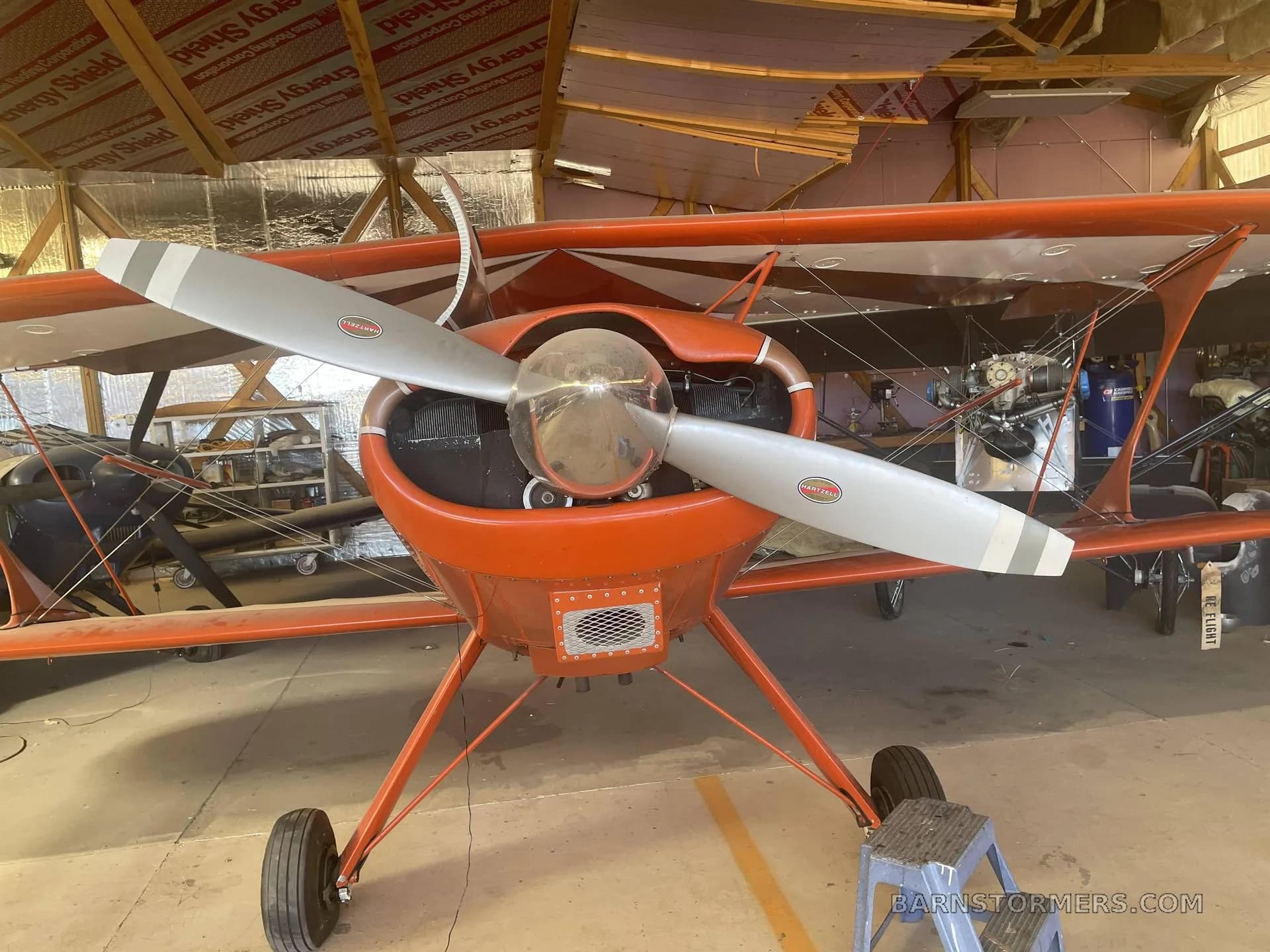 A front view of an orange and white Star Duster Too biplane with a large propeller, parked in a hangar.