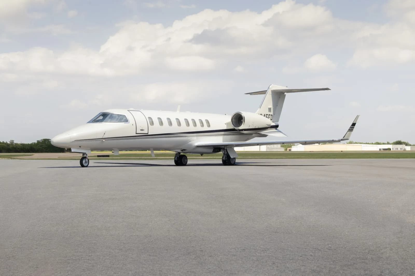 A white Learjet 45 business jet with black accent stripes is parked on an asphalt tarmac under a partly cloudy sky.