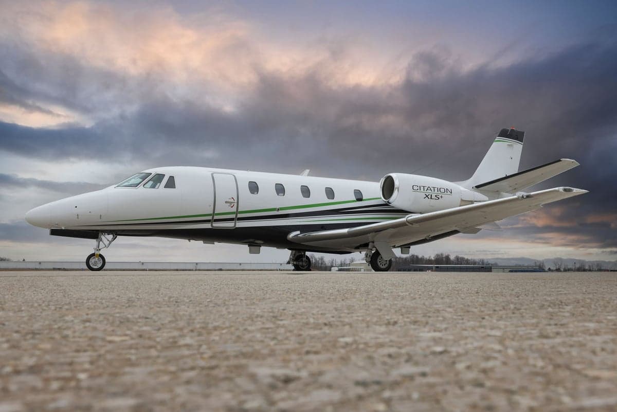 A white Cessna Citation XLS+ business jet with green and black stripes parked on a concrete tarmac under a dramatic cloudy sky.