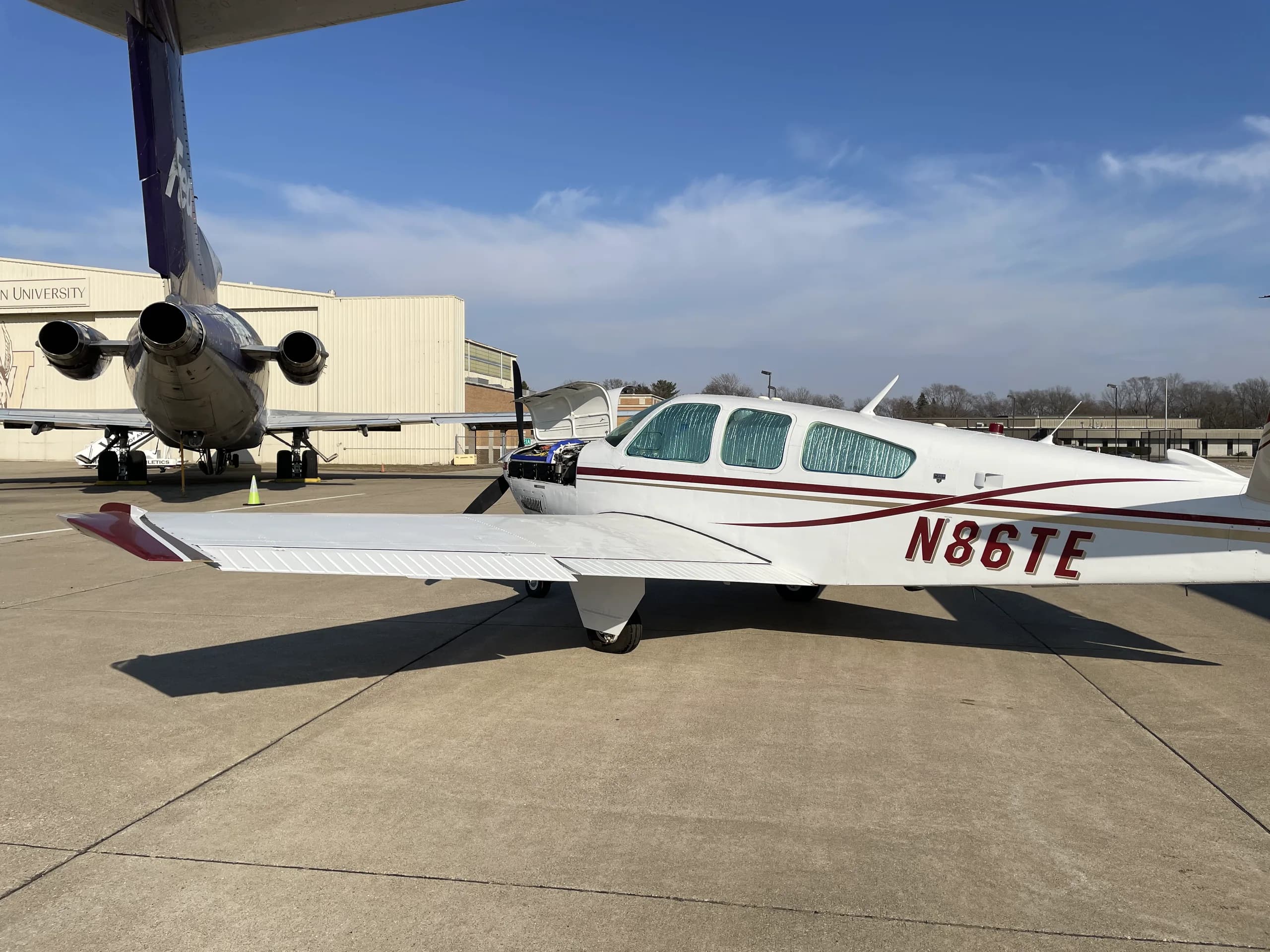 Side view of a white Beechcraft Bonanza, N86TE, with red and gold stripes, parked on an airfield next to a jet.