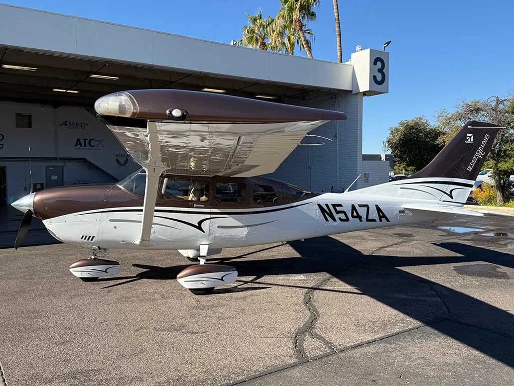 A brown and white Cessna 182 Skylane N54ZA with a high wing and fixed gear is parked outside a hangar on a sunny day.
