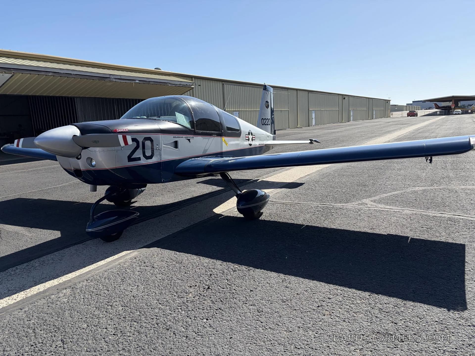 A front-quarter view of a silver and blue Grumman American AA-5 Traveler aircraft with '20' on the nose, parked on an airfield.