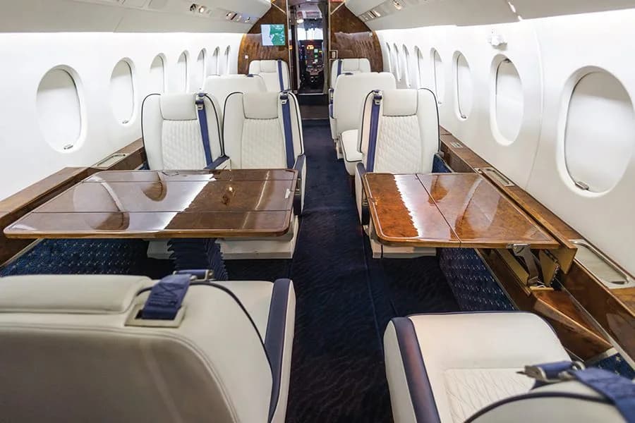 The interior of a private jet cabin with white and blue seats and two polished wooden tables.