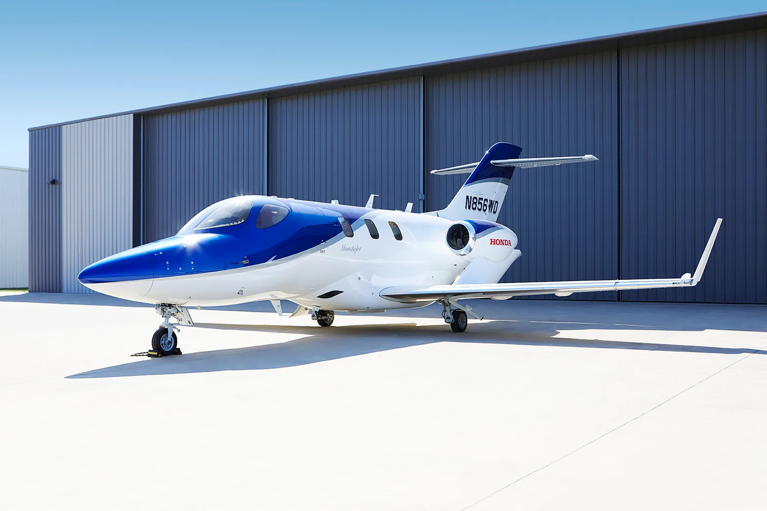 Three-quarter rear view of a blue and white HondaJet N856WD parked on an airfield in front of a hangar.