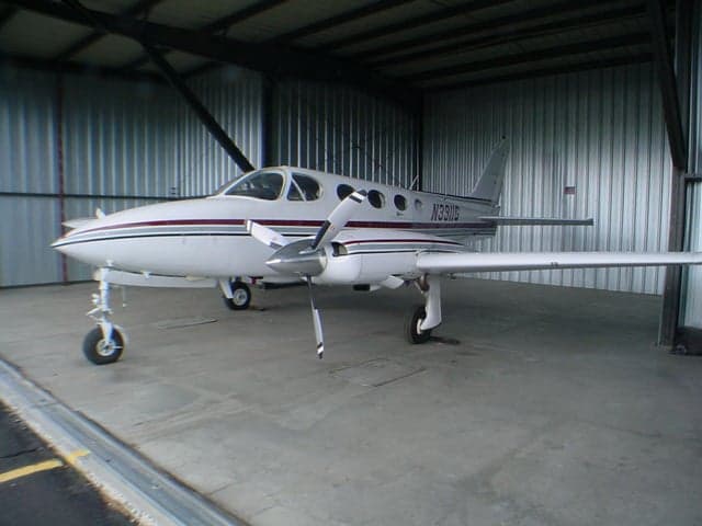 A front-quarter view of a white twin-engine aircraft with red and grey stripes, registration N3911G, parked inside a metal hangar.