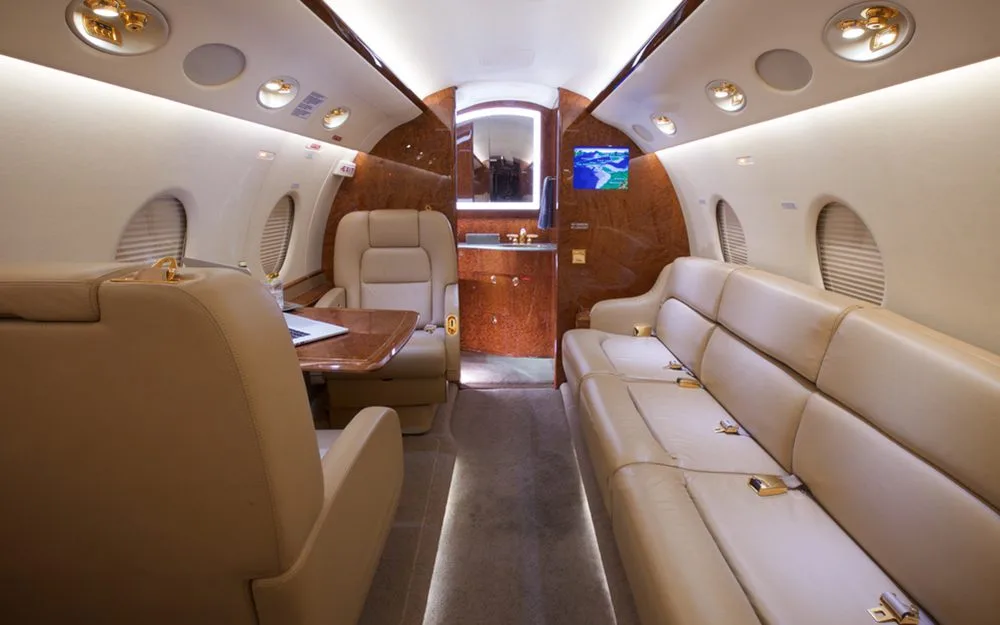 A luxurious aircraft cabin with a tan leather sofa, individual seats, and a view towards the rear lavatory/galley area.