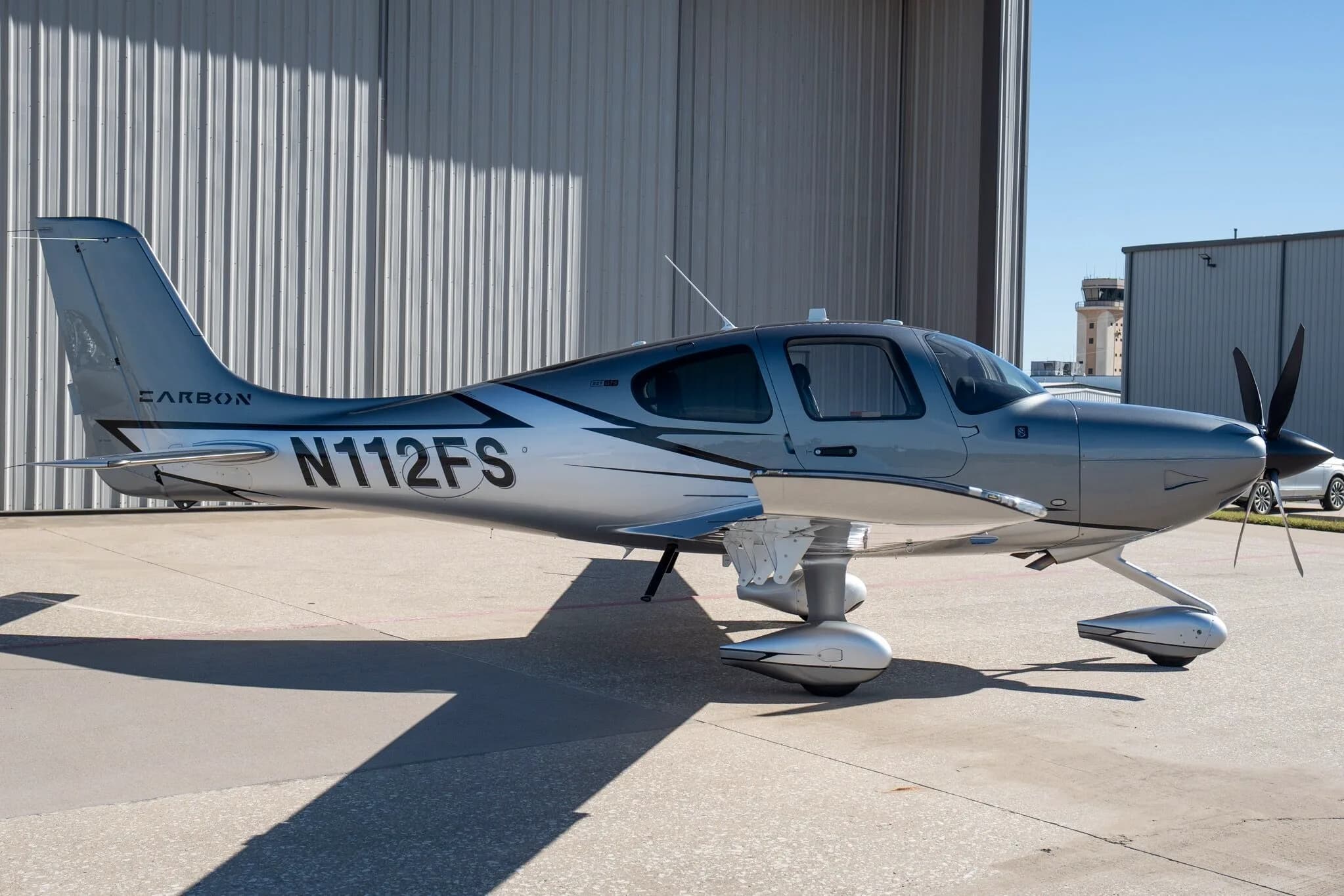 A side view of a silver and dark grey Cirrus SR22T Carbon Edition aircraft (N112FS) on an airport tarmac with a control tower in the distance.