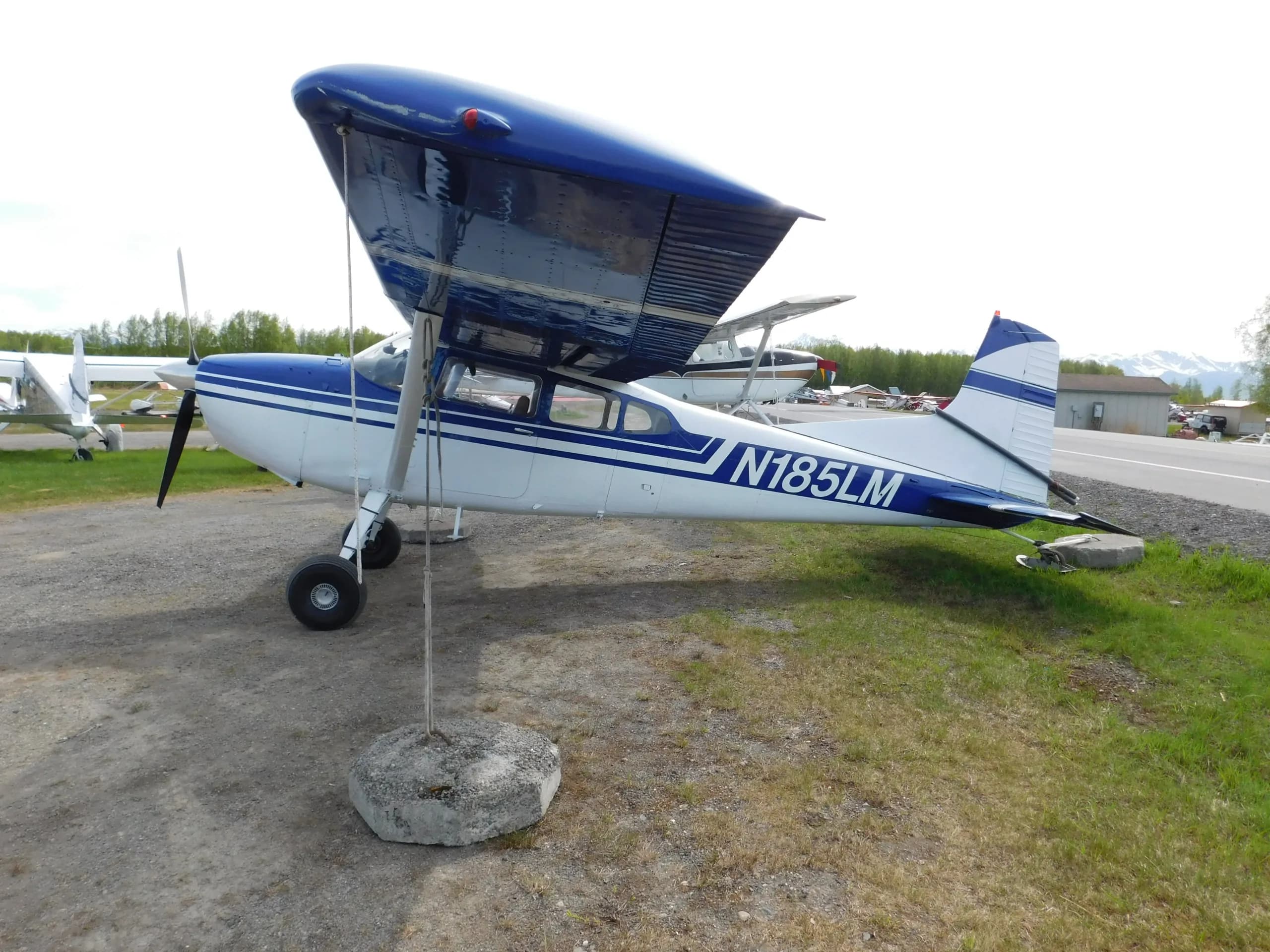 A white and blue Cessna 185 aircraft with registration N185LM is parked on a gravel and grass airfield.