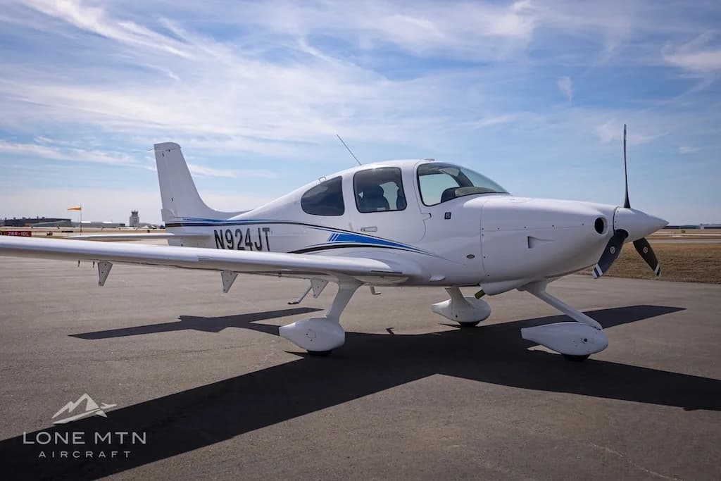 A white single-engine aircraft with blue and black stripes, tail number N924JT, parked on an airport tarmac under a partly cloudy sky.