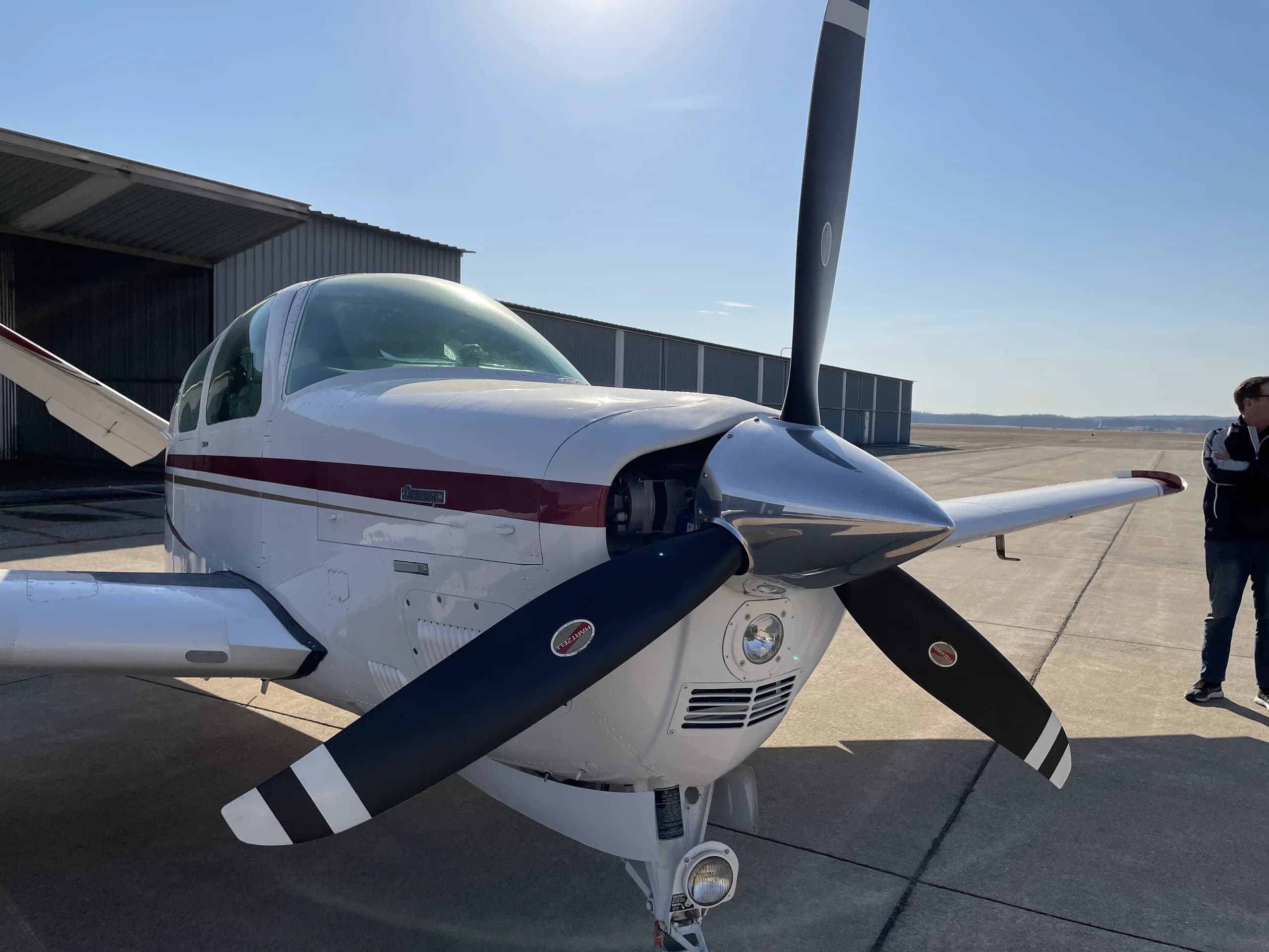 A white single-engine propeller aircraft with a red stripe, parked on an airfield with a hangar in the background.