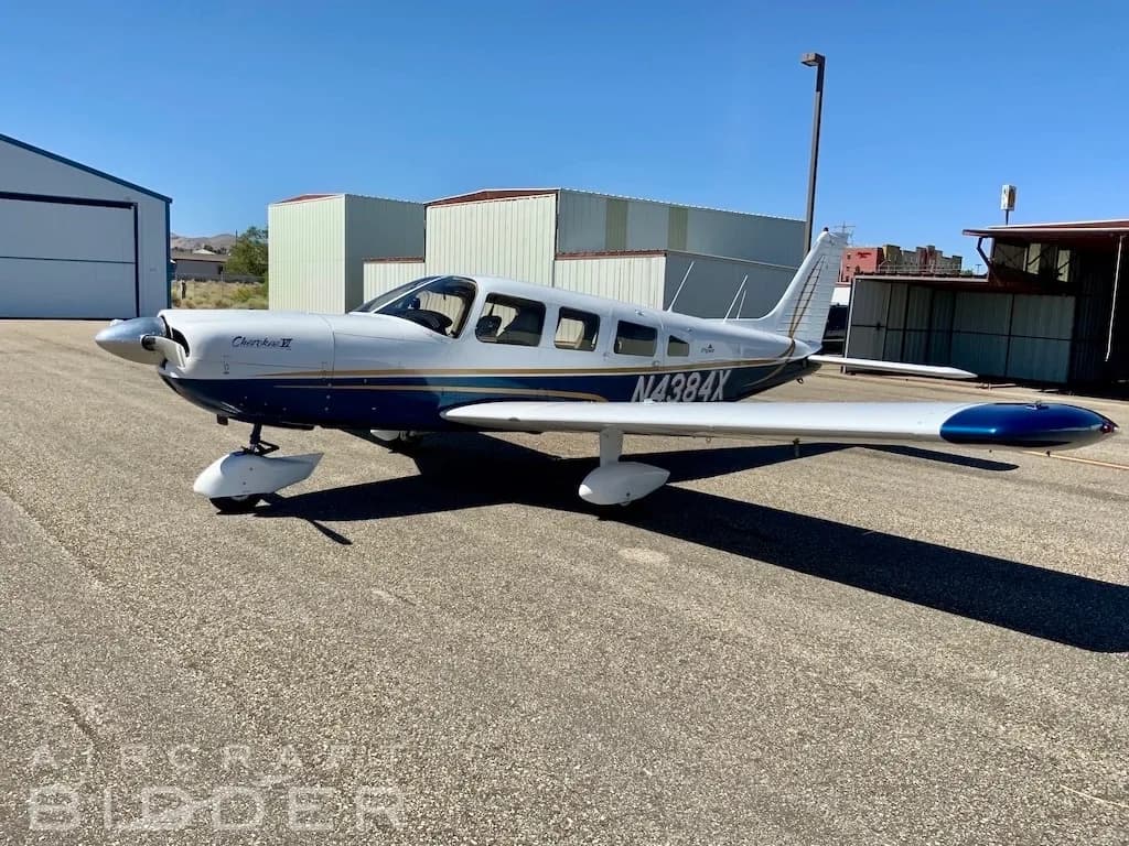 A white and blue Piper Cherokee VI aircraft, registration N4384X, parked on an asphalt ramp in front of hangars under a clear sky.