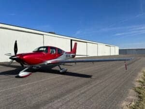 A Cirrus SR22T aircraft with a red and silver paint scheme is parked on a tarmac in front of a hangar.