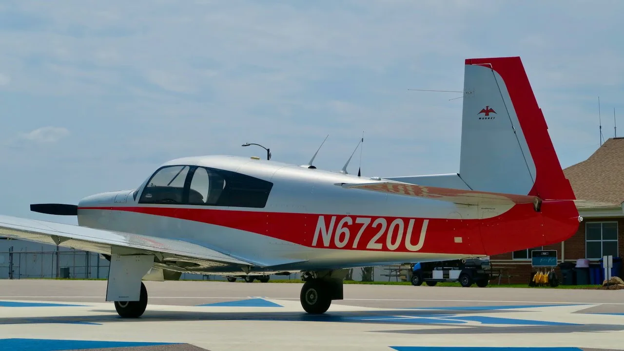 Left side view of a silver and red Mooney aircraft with registration N6720U parked near a building.
