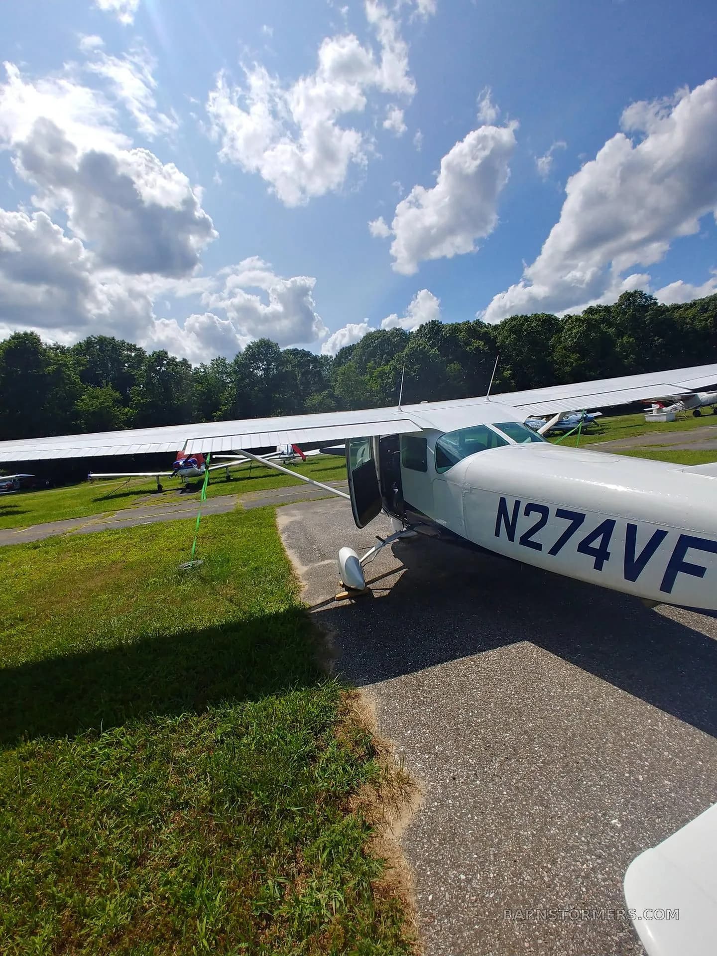 Three-quarter front view of a white and blue single-engine aircraft (N274VF) with its door open, parked on an asphalt path.