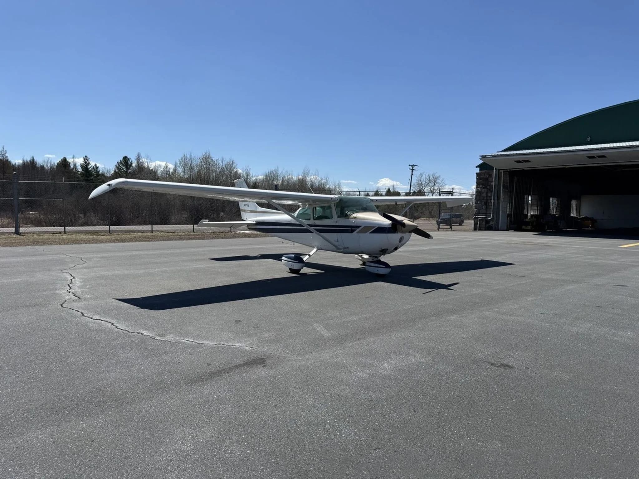A white and tan Cessna 172 aircraft with blue stripes parked on an asphalt apron next to a green hangar.