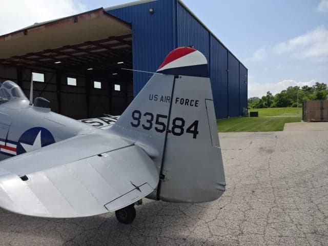 Close-up of the tail of the silver T-6 Texan aircraft showing the US Air Force marking and tail number, with a hangar in the background.