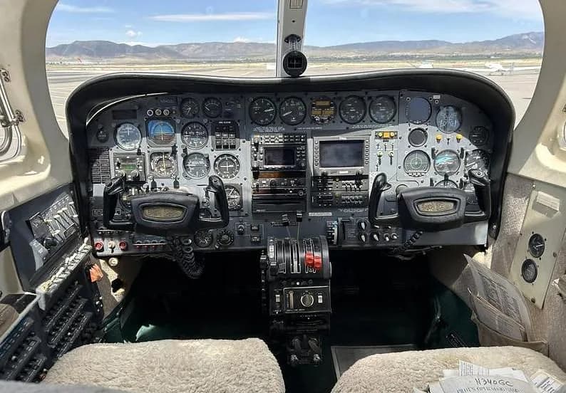 The cockpit of a Cessna 340A, showing the instrument panel, yokes, and throttle quadrant, with an airport runway visible through the windshield.