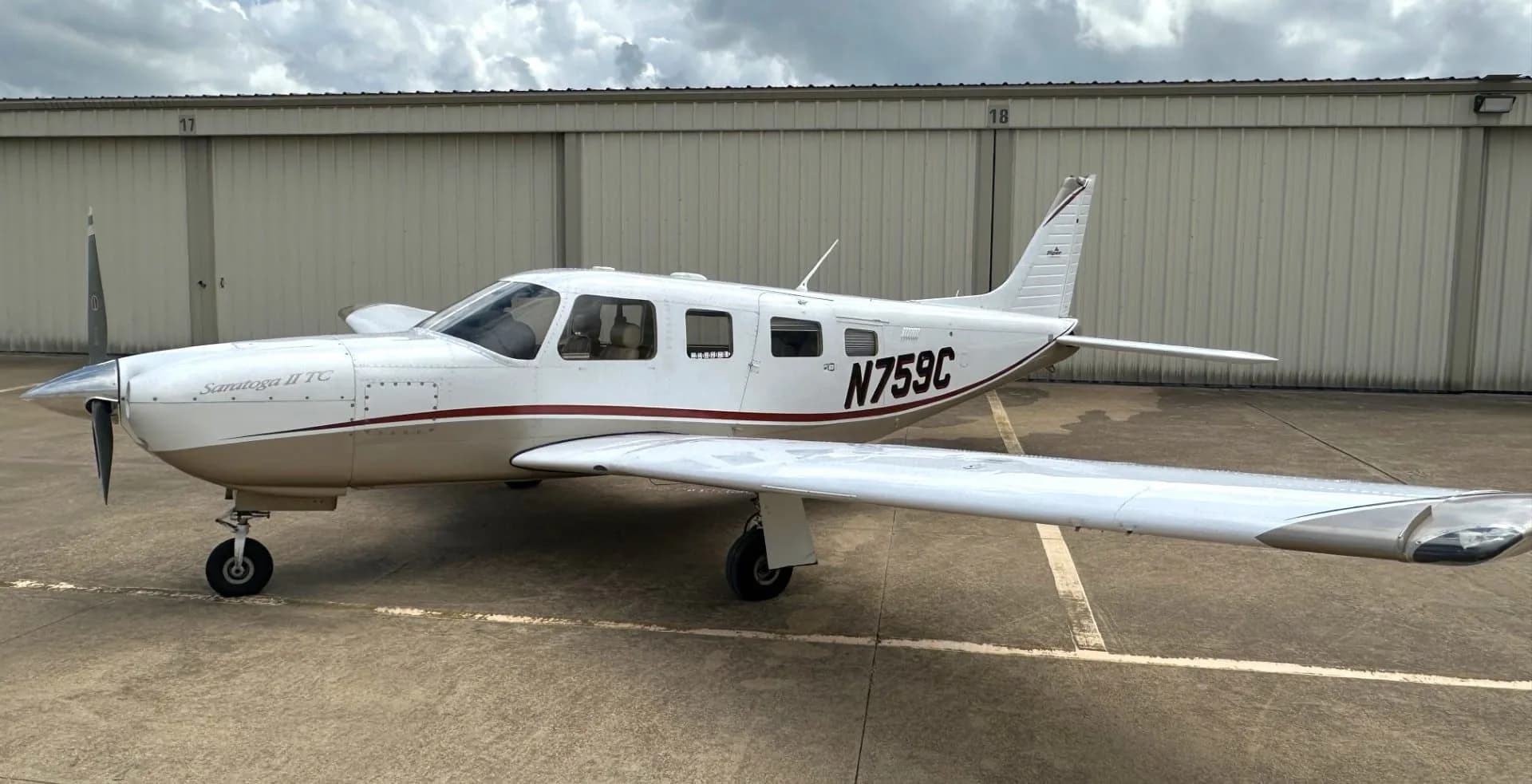 A white and tan Piper Saratoga II TC aircraft (N759C) parked on an asphalt ramp in front of hangars under a cloudy sky.