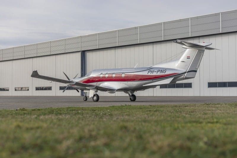 A Pilatus PC-12 aircraft with a silver and red paint scheme, registration PH-PNG, parked on the tarmac in front of a hangar.