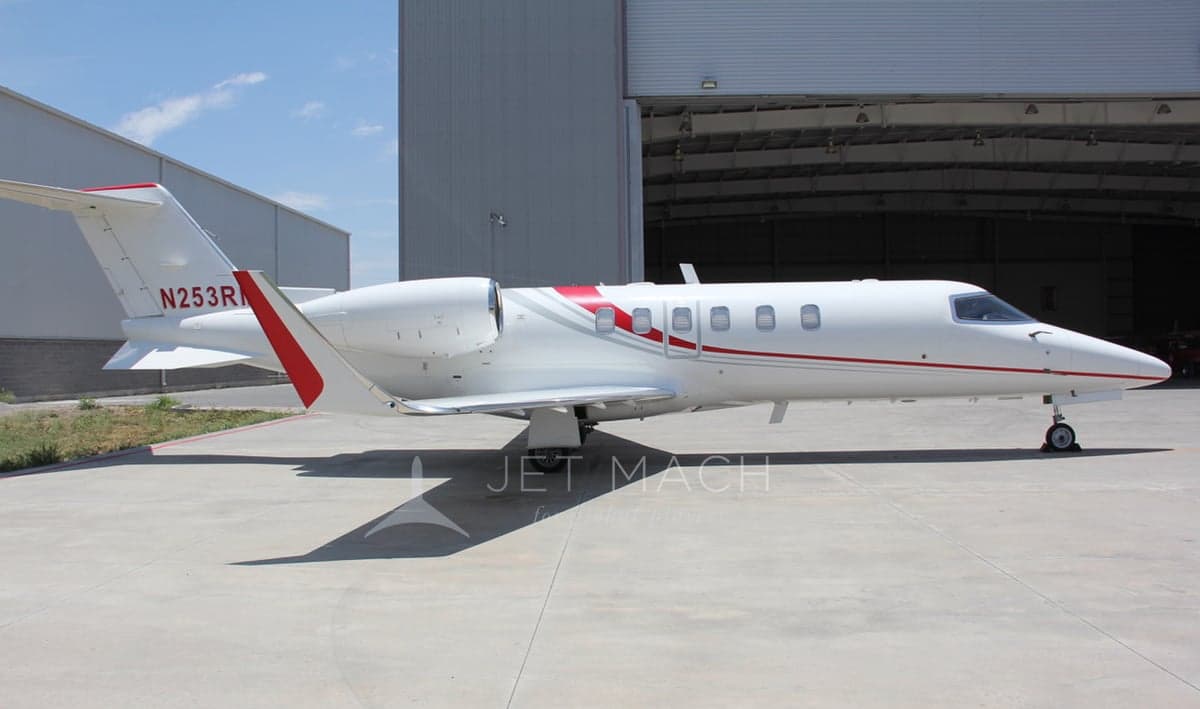 A white Learjet 75 with red and grey stripes, registration N253RM, parked on a concrete apron outside a hangar.