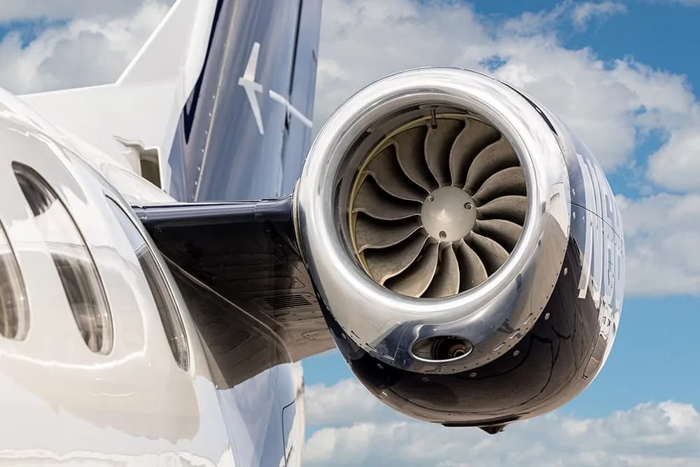 A close-up view of the polished chrome engine nacelle and fan blades of the Embraer Phenom 100E, N68NH, against a cloudy sky.