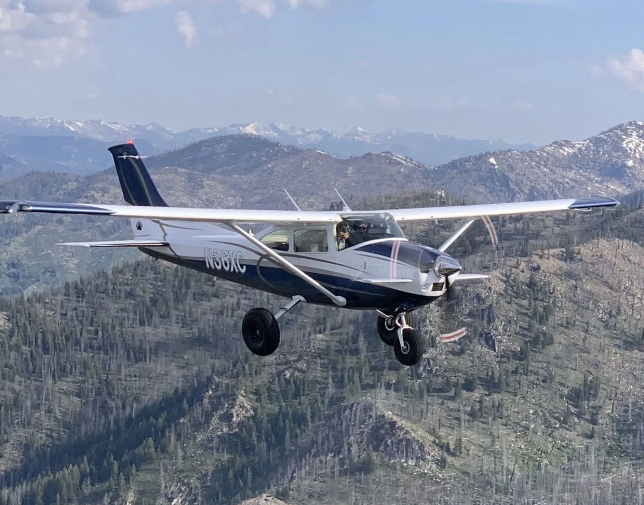 A white and blue Cessna 182 aircraft with registration N36XC flying over a mountainous, forested landscape.