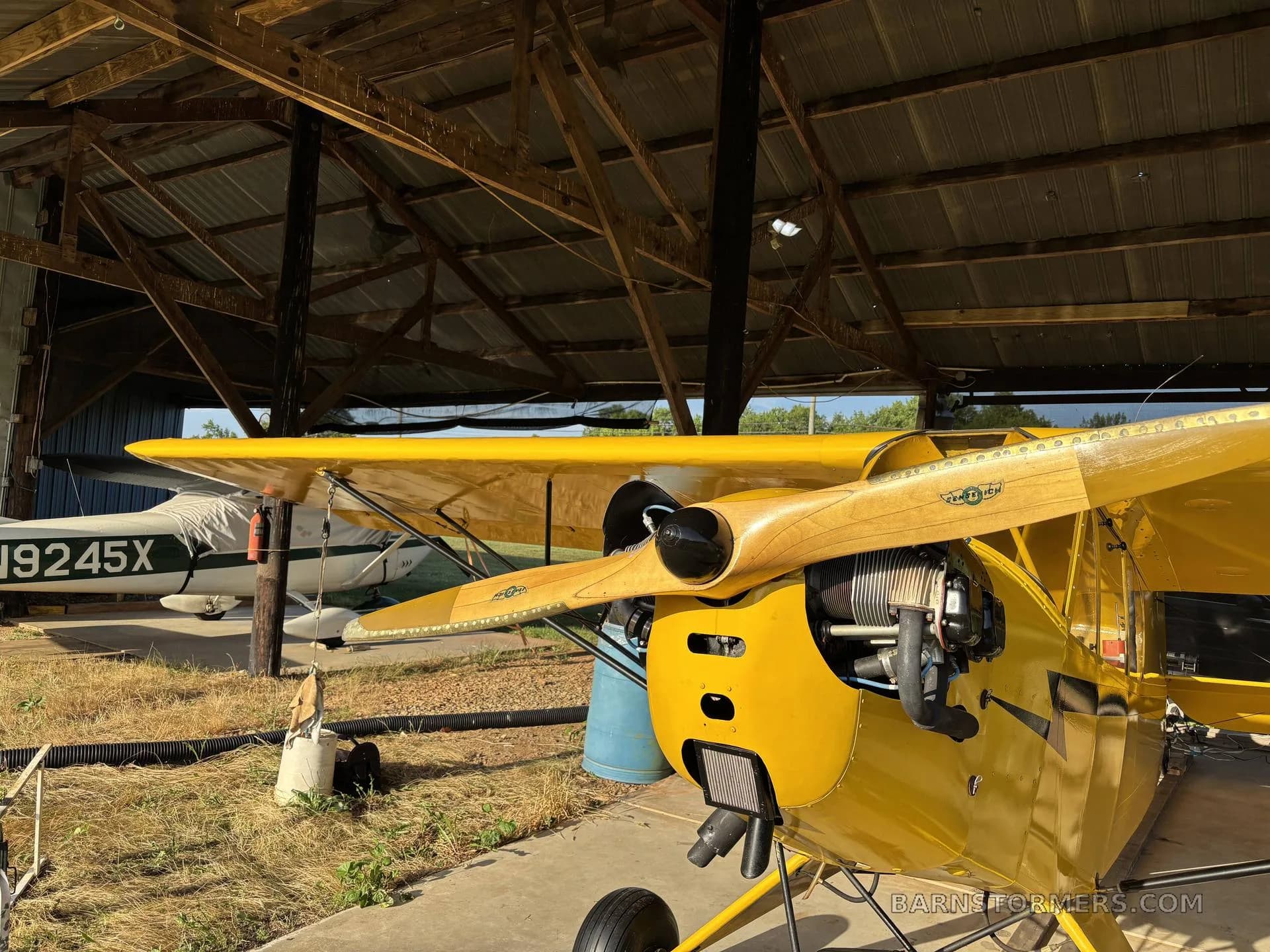 The front view of a yellow aircraft with a wooden propeller, parked in a hangar next to a white aircraft.
