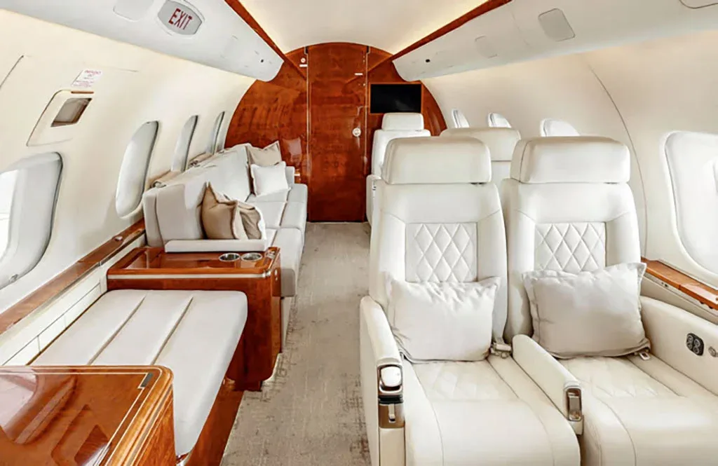 The spacious private jet cabin features white leather seats, a lounge sofa, and polished wood accents.