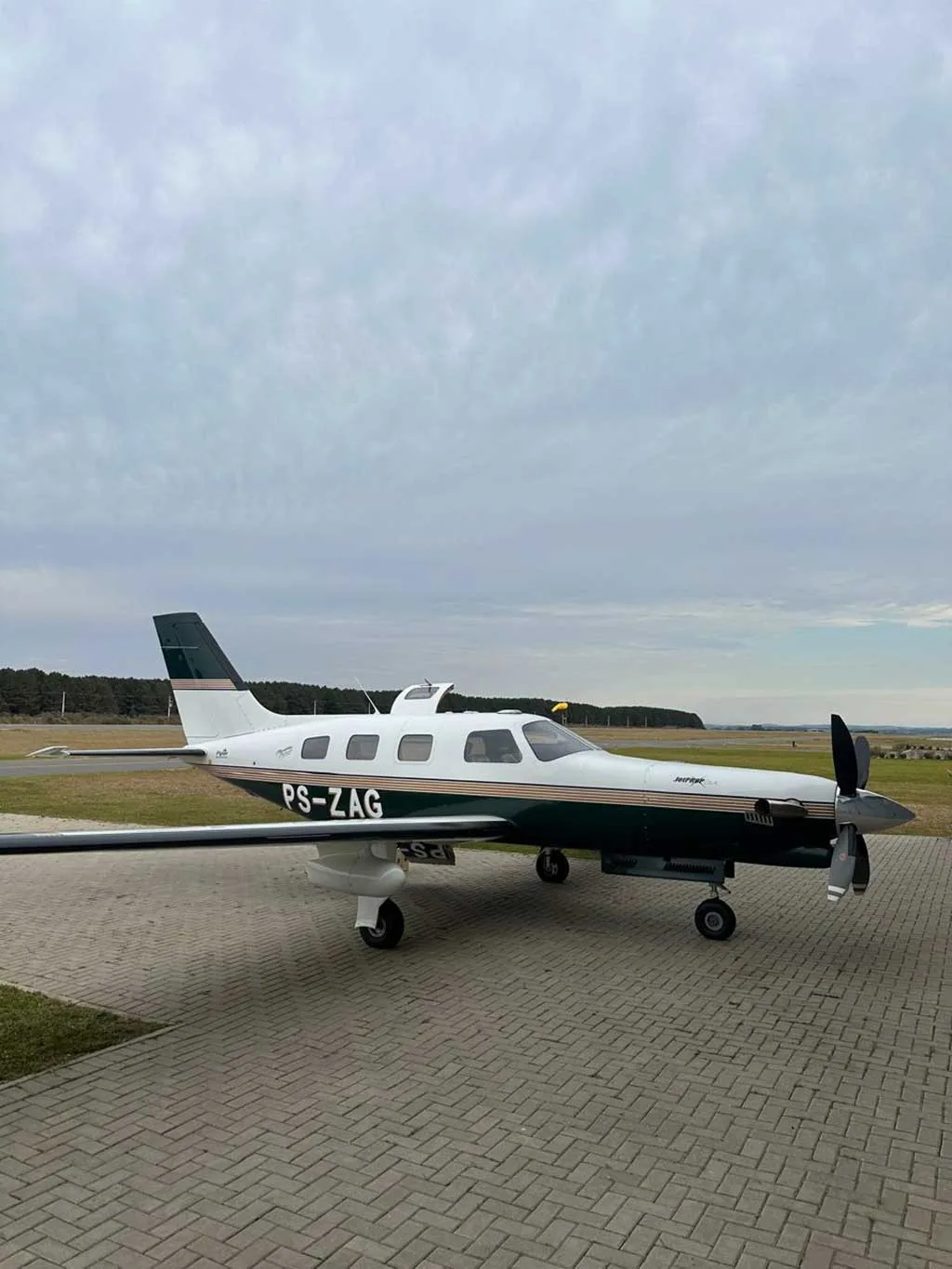 A white and dark green single-engine turboprop aircraft, PS-ZAG, parked on a paved apron under a cloudy sky.