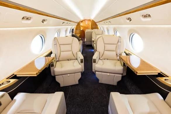 The luxurious interior of a private jet cabin with beige leather seats, wooden tables, and round windows.