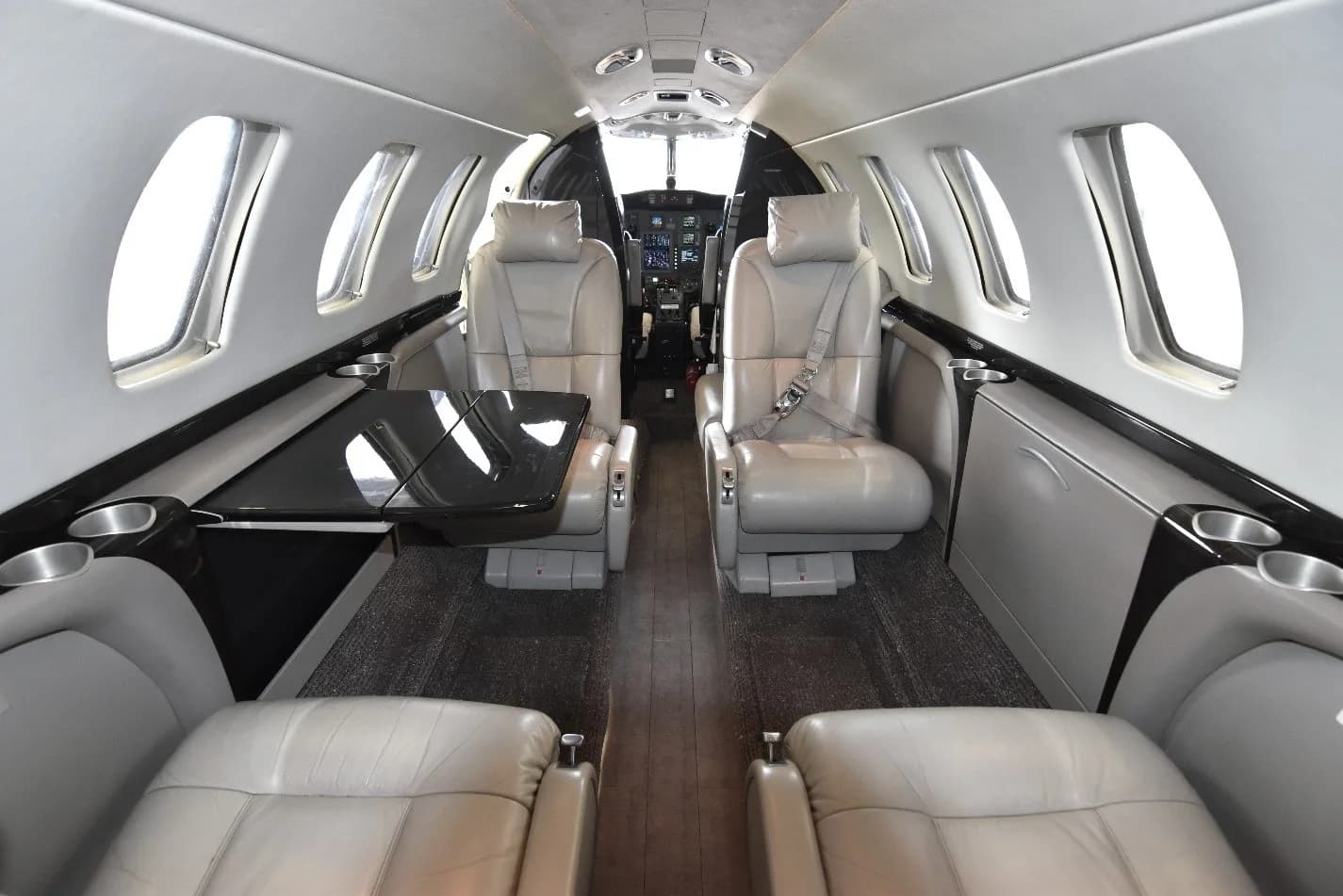 The interior cabin of a private jet, showing light gray leather seats, a dark table, and the cockpit in the background.
