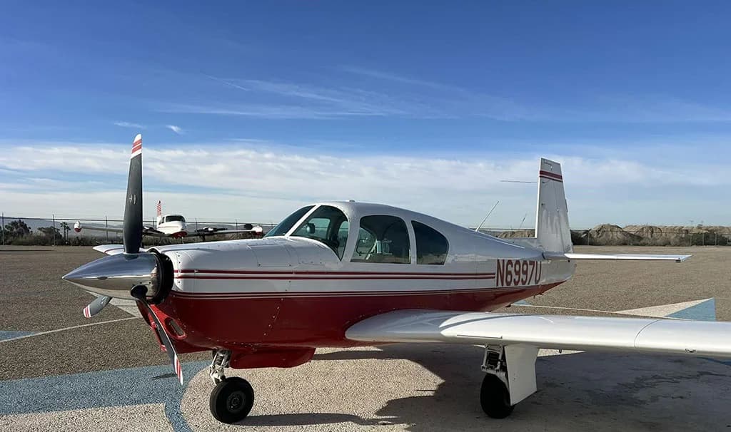 A white and red single-engine aircraft, N6997U, with a propeller, parked on an airfield under a blue sky.