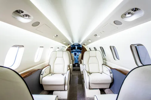 The interior of an aircraft cabin with rows of cream-colored leather seats and overhead lighting, looking towards the cockpit.