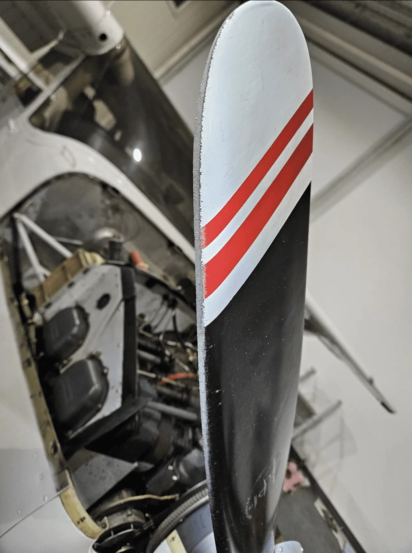 A close-up of an aircraft propeller with black and white blades featuring red stripes, with the engine compartment visible behind it.
