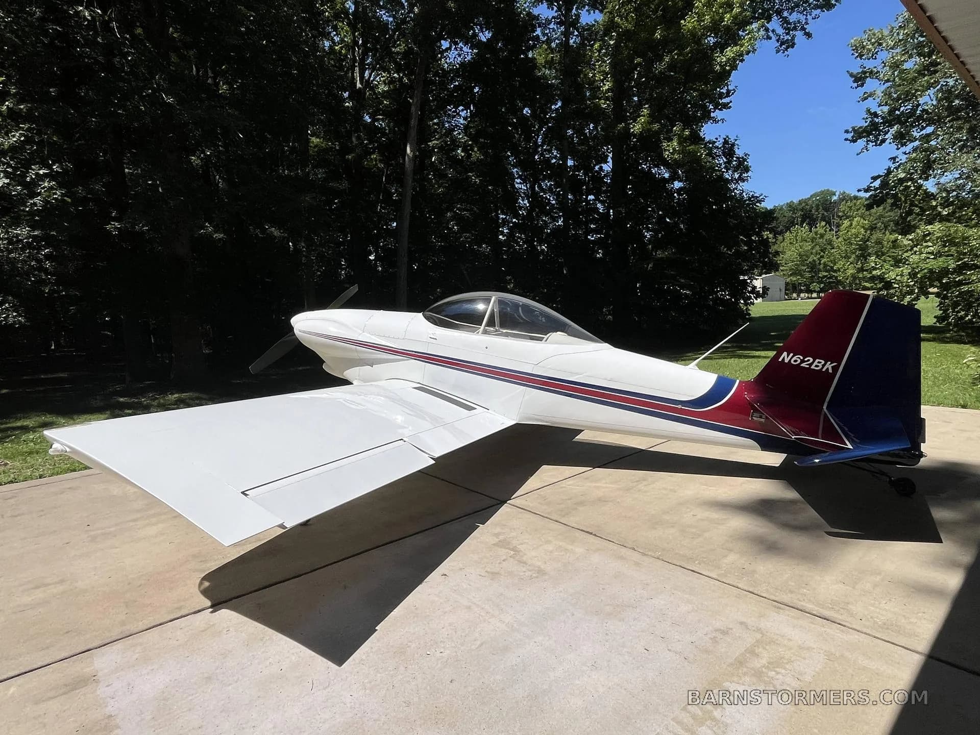 A white single-engine aircraft with red and blue tail stripes, viewed from the side, parked on concrete with trees in the background.