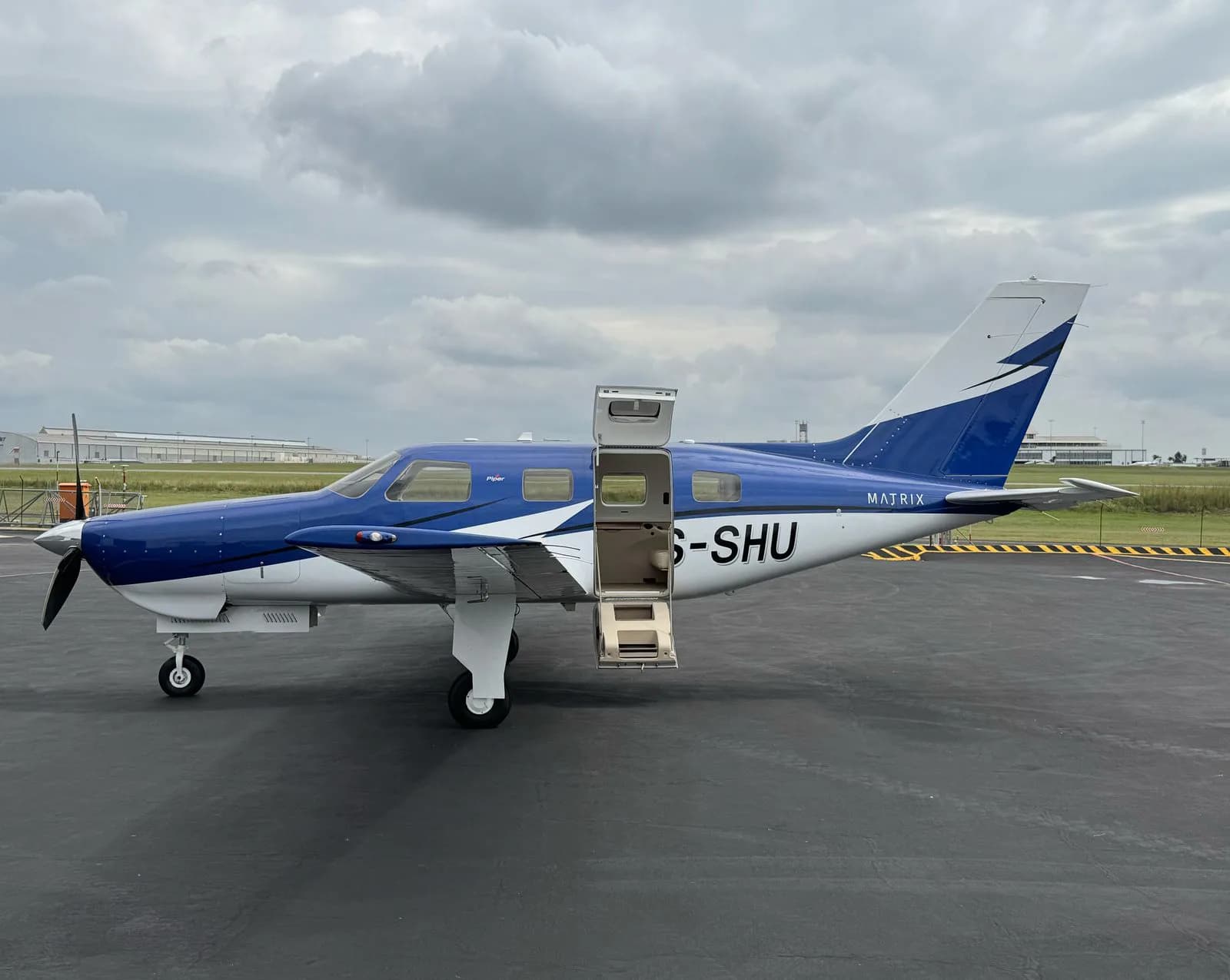 A blue and white Piper M600 Matrix aircraft with registration G-SHU parked on an tarmac with its door open.