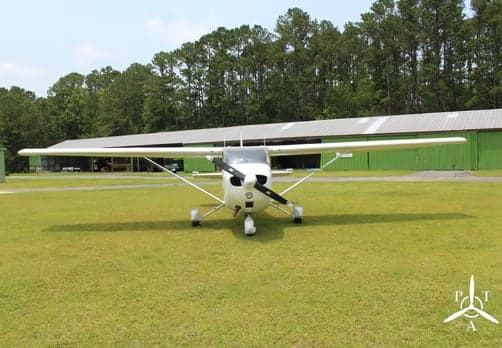 A white Cessna 172 aircraft with a black propeller is viewed head-on, parked on a grassy airfield.