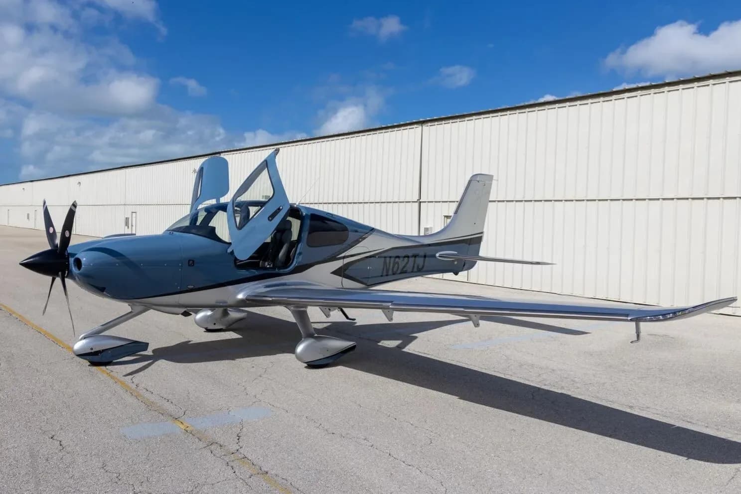 A three-quarter front view of a blue and silver Cirrus SR22T aircraft with its gull-wing doors open, parked on an airfield.