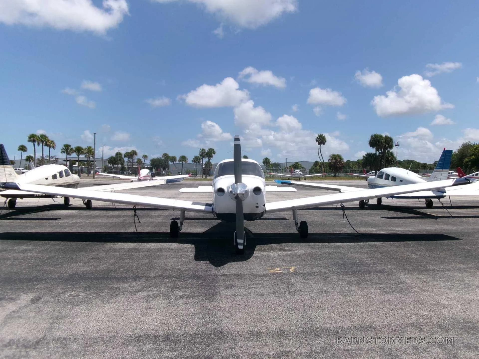 A front view of a white Piper PA-28 Cherokee with a black propeller, parked on an asphalt ramp surrounded by other small aircraft and palm trees.